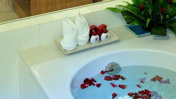 A bathroom with a bathtub filled with rose petals, a towel rack with folded towels, a small plant, and a bedside table with a lamp and a telephone.
