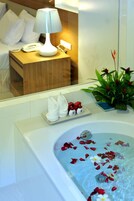 A bathroom with a bathtub filled with rose petals, a towel rack with folded towels, a small plant, and a bedside table with a lamp and a telephone.