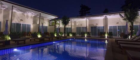 A swimming pool with lounge chairs and umbrellas, surrounded by a building with large windows and plants.