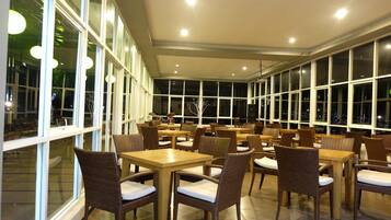A spacious indoor dining area with multiple tables and wicker chairs, large windows providing views of the night sky.
