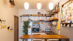 A compact kitchen with a wooden table, white chairs, and a variety of kitchen appliances. There are hanging lights, a potted plant, and a wall with notes and pictures. - Restiny Hostel (Bangkok)