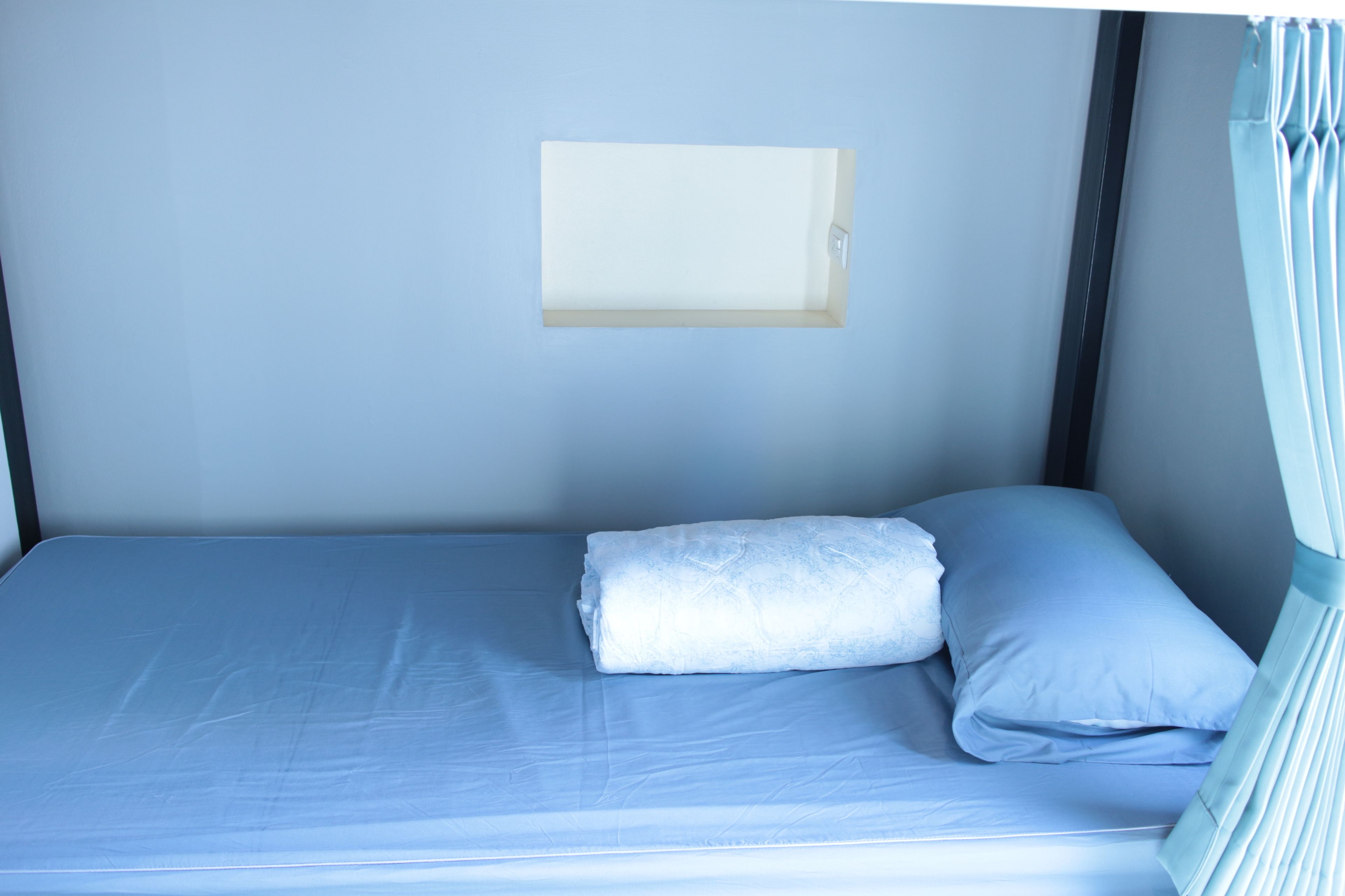 A single bed with a blue mattress and pillows, a small bedside table, and a wall-mounted light fixture.