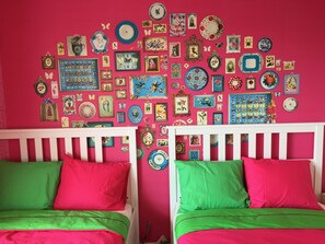 Habitación con dos camas, cada una con ropa de cama verde y rosa, y una pared rosa vibrante decorada con numerosas fotos enmarcadas y platos decorativos.