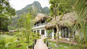 A bungalow with a thatched roof, surrounded by lush greenery and a paved pathway leading to the entrance. - Tam Coc Rice Fields Resort (Nam Hoa Lu)