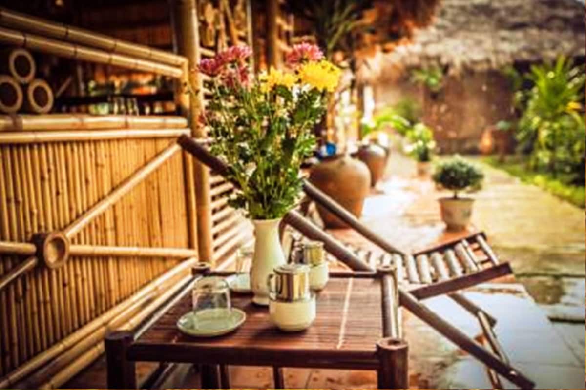 A bamboo structure with a table set for tea, surrounded by potted plants and a thatched roof.