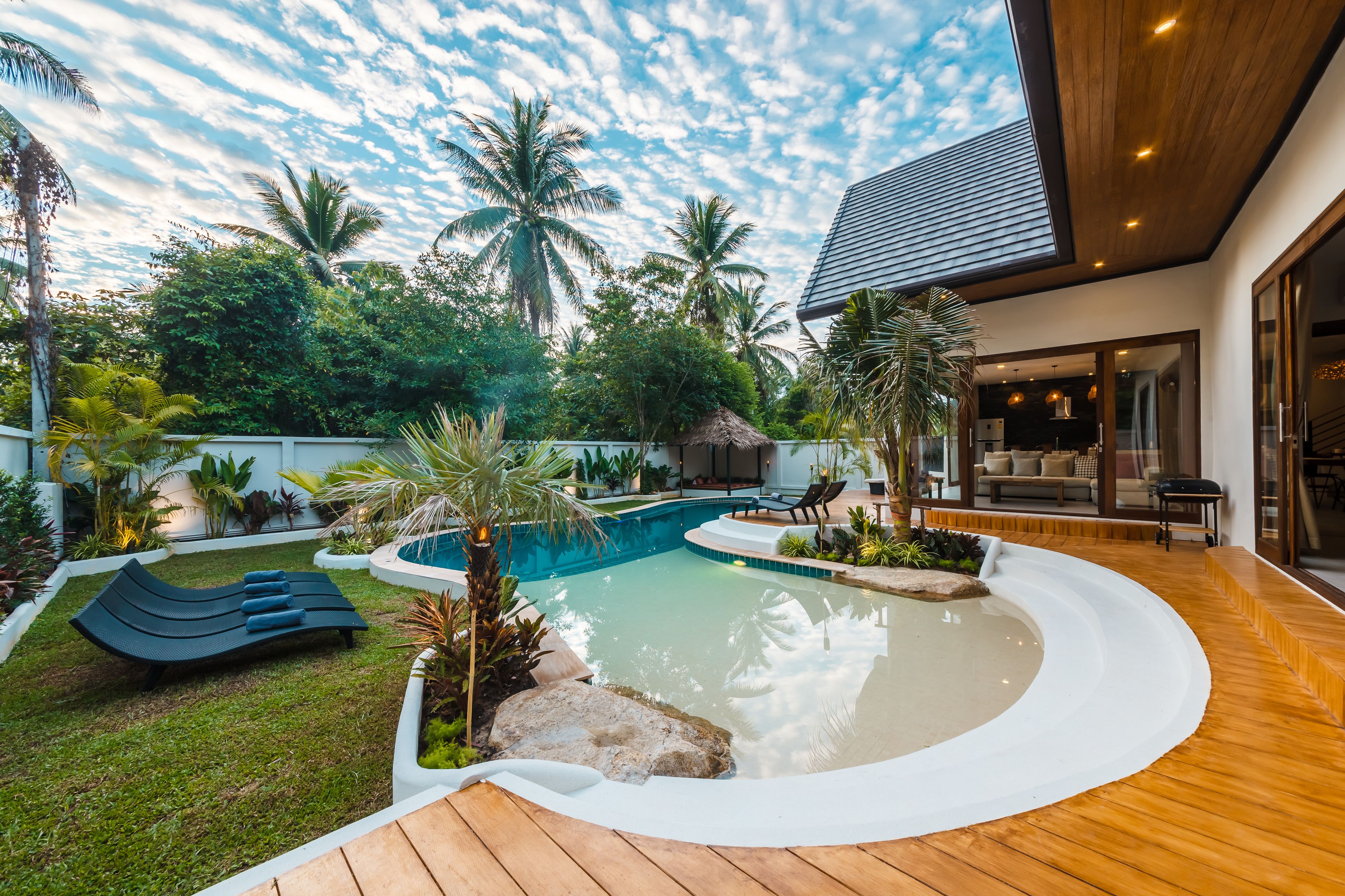 A modern outdoor pool area with lounge chairs, a wooden deck, and tropical plants.