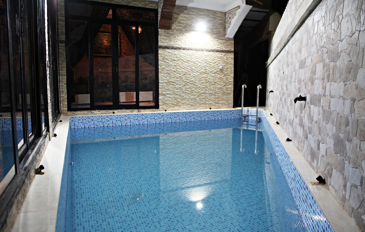 Ein Swimmingpool mit blauen Mosaikfliesen, einer Steinmauer an einer Seite und einer Holztür mit Glasscheiben.