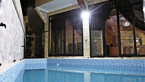 An indoor swimming pool with a tiled edge, large windows with black frames, and a visible staircase in the background.
