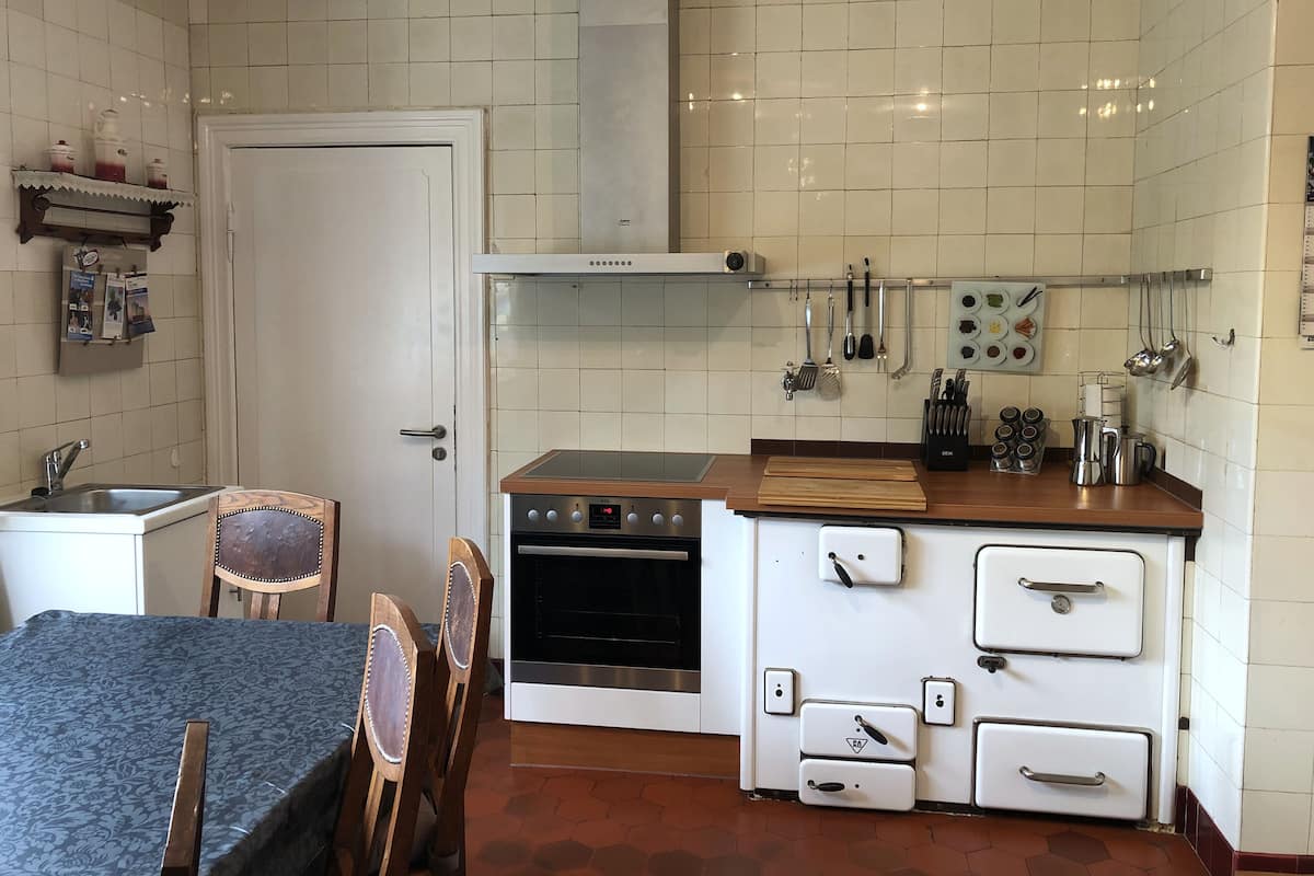A compact kitchen with white tiled walls, a stove, oven, and a small sink. There are wooden chairs and a table with a blue tablecloth.