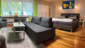 Una sala de estar moderna con un sofá gris, una mesita con una botella y vasos, una cama con ropa de cama gris y una obra de arte azul en la pared.