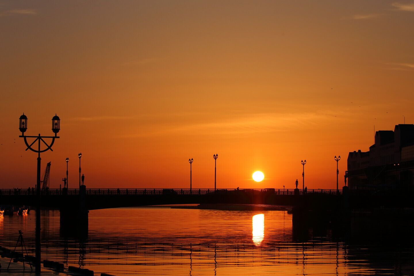 A sunset over a waterfront with a bridge, street lamps, and a statue.