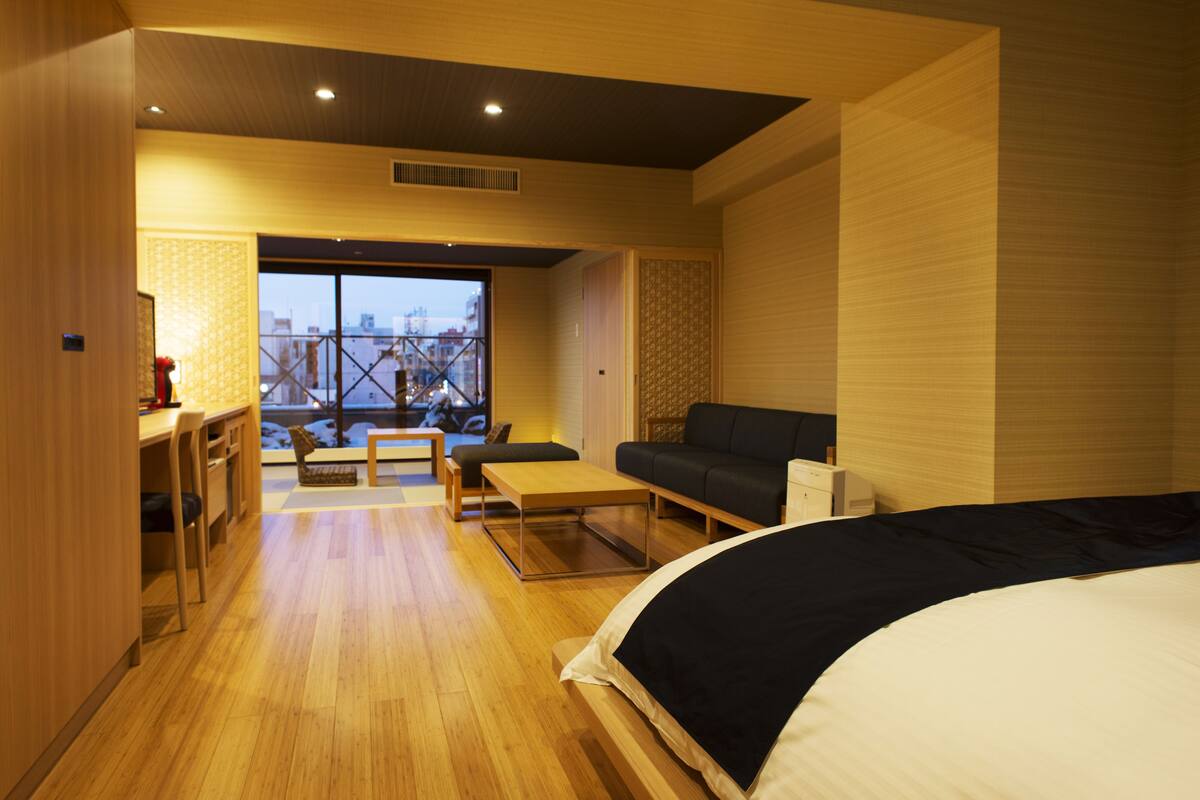 A modern hotel room with a large bed, a sofa, a dining table, and a view of the cityscape through a large window.