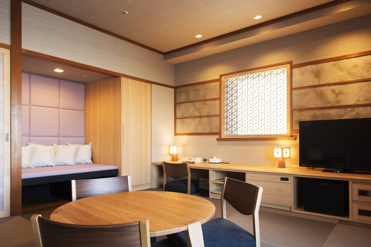 A modern hotel room with a wooden dining table, chairs, a sofa with cushions, a television, and a desk with a lamp.