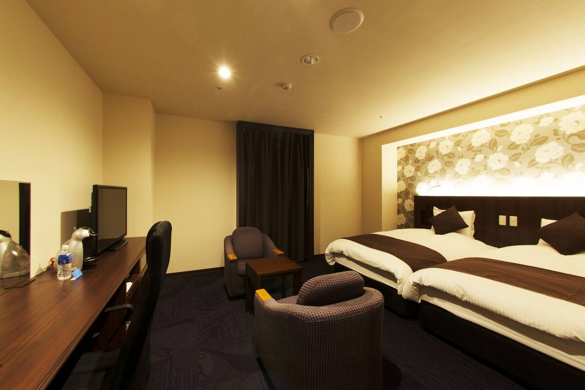 A hotel room with a desk, two beds, a chair, a TV, and a window with curtains.