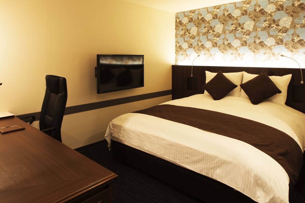 A hotel room with a bed, a TV mounted on the wall, a desk, and a chair.