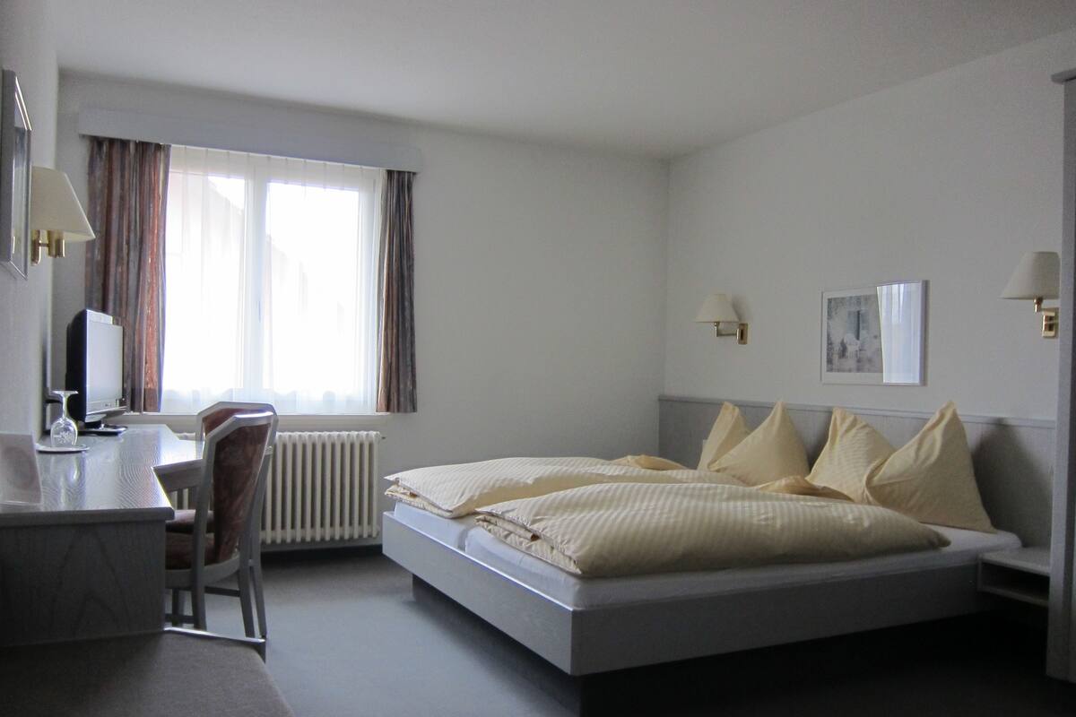 A hotel room with a bed, a desk, a chair, a radiator, and a window with curtains.