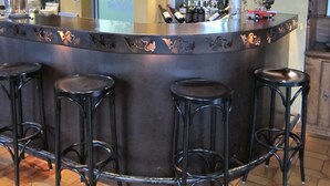 Zona de bar con mostrador curvo, varios taburetes y una amplia selección de botellas y vasos.