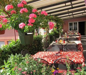 An outdoor dining area with tables covered in red floral tablecloths, surrounded by lush greenery and pink flowers. - Landhotel Hirschen (Truellikon)