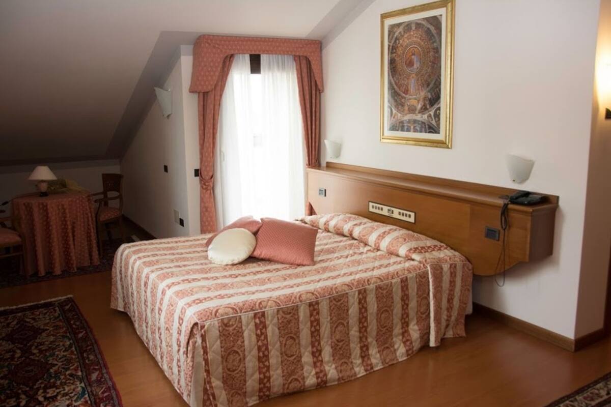 A hotel room with a bed, a desk, a chair, a lamp, a painting, and a window with curtains.