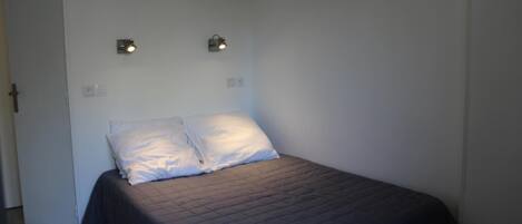 Una cama individual con una colcha azul oscuro y dos almohadas, dos luces fijadas a la pared y una puerta blanca.