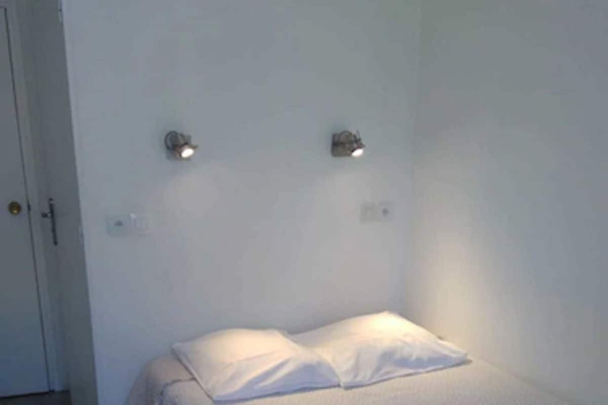 Uma cama pequena e bem arrumada, com um edredom branco e almofadas, um tapete escuro e uma porta branca.