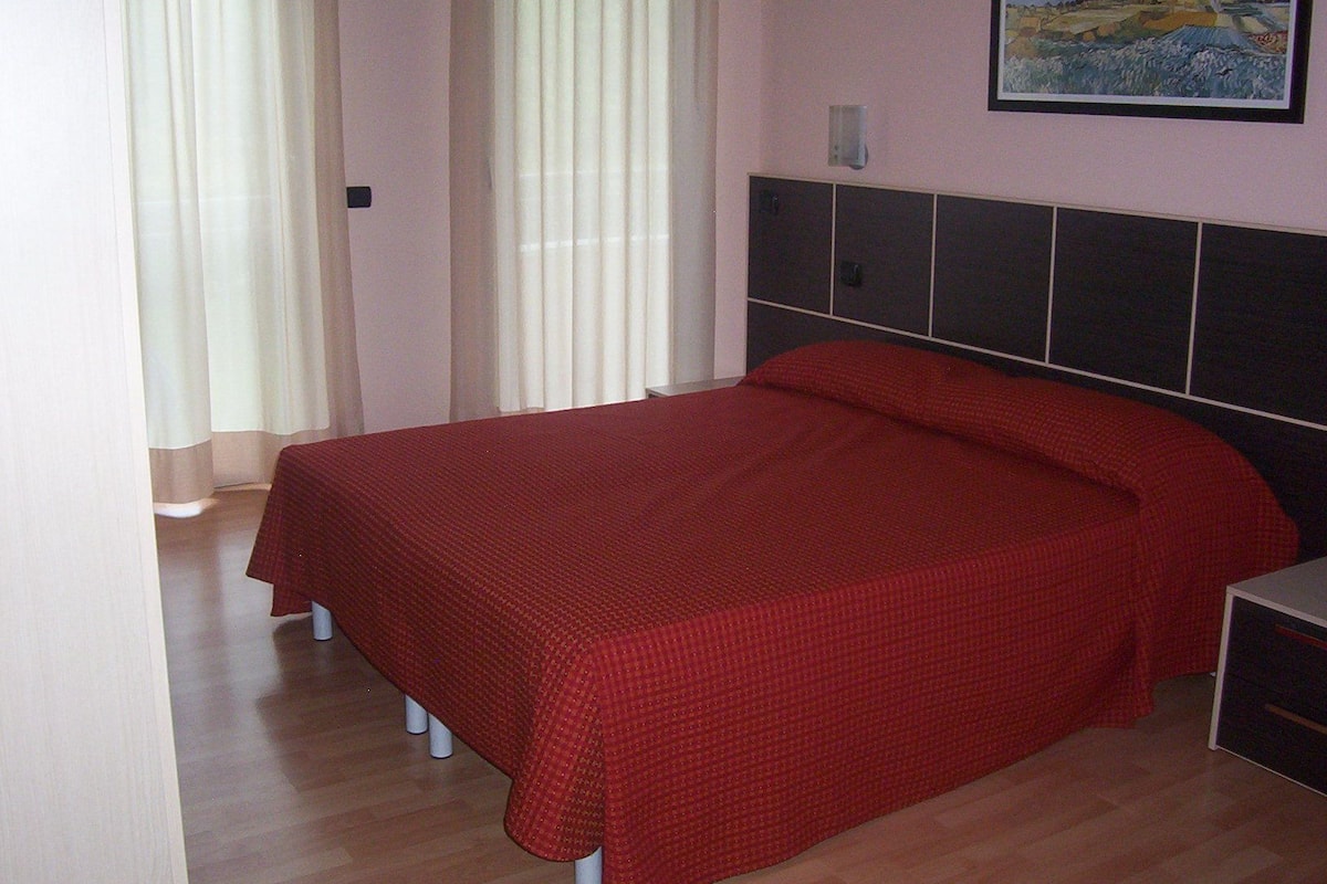 A bedroom with a bed, a nightstand, a painting on the wall, and a window with curtains.