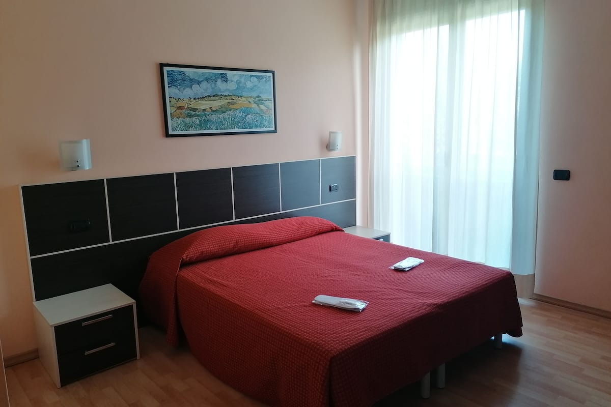 A hotel room with a bed, a nightstand, a wall-mounted light, a painting, and a window with curtains.