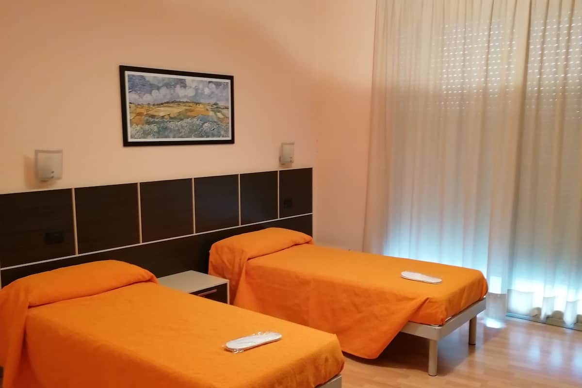 A hotel room with two single beds, a headboard, a painting, a window with curtains, and a wooden floor.