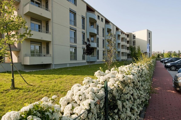 A multi-story residential building with balconies, a paved driveway, and a row of parked cars.