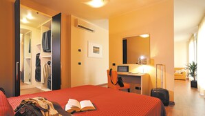A hotel room with a bed, a desk with a computer, a chair, a suitcase, and a wardrobe.