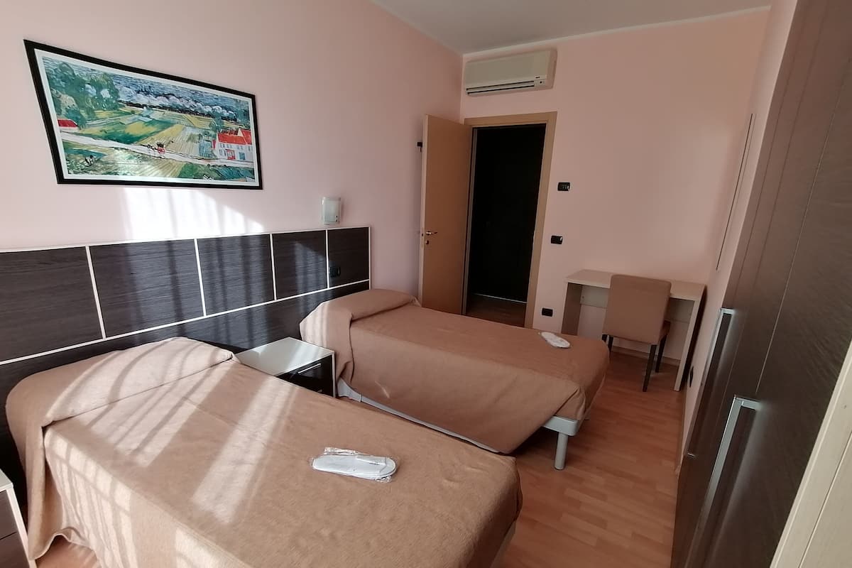 A hotel room with two beds, a desk, a chair, an air conditioning unit, and a painting on the wall.