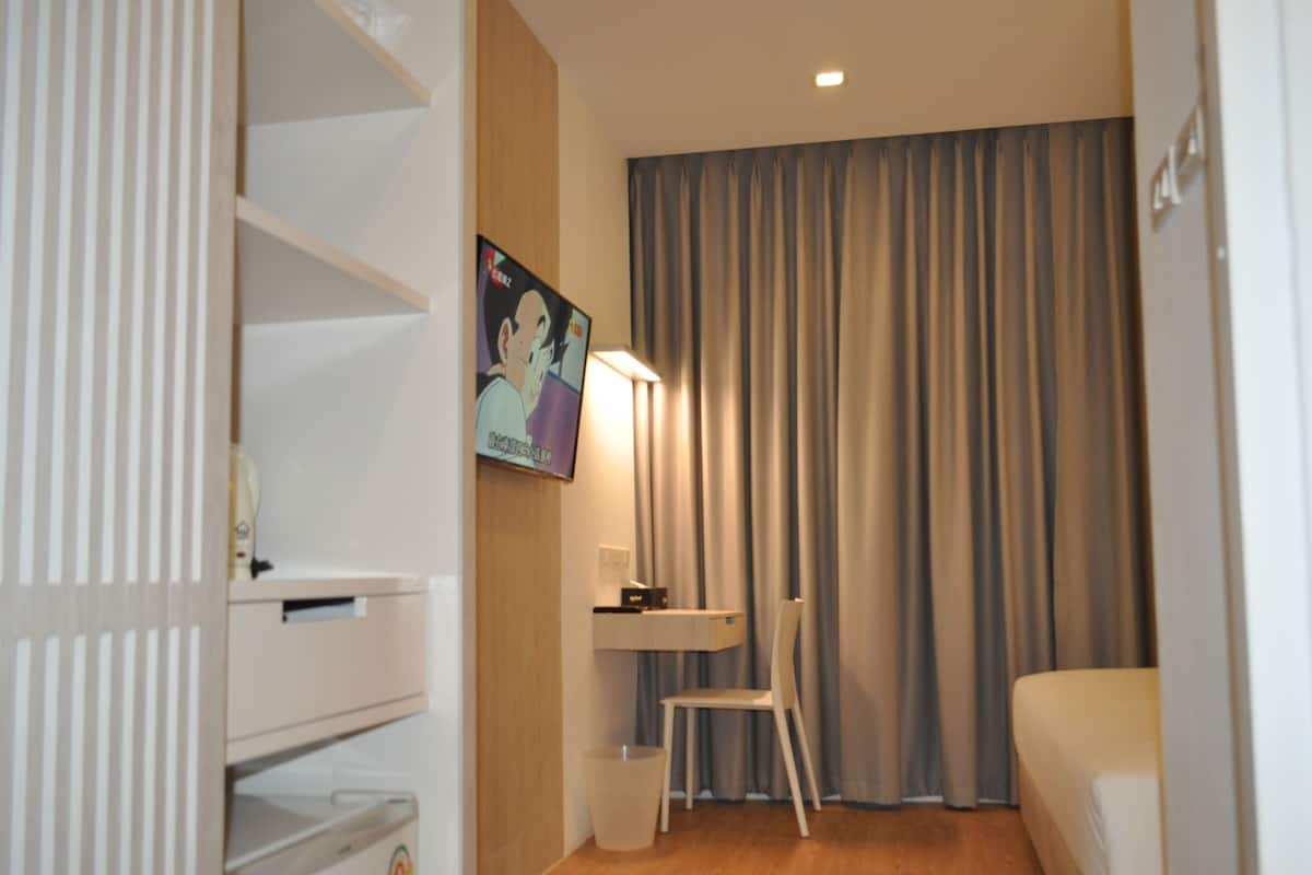 Una habitación de hotel con una cama, un escritorio con una silla, una mesita pequeña, un cubo de basura y un cuadro en la pared.