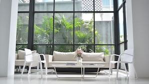 Ein modernes Wohnzimmer mit einem weißen Sofa, zwei weißen Sesseln, einem Glastisch und einer Vase mit Blumen. Große Fenster bieten Blick auf Grünflächen außerhalb.
