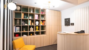 A reception area with a wooden reception desk, a yellow chair, and a bookshelf filled with books and decorative items. - Hotel Esperance (Paris)
