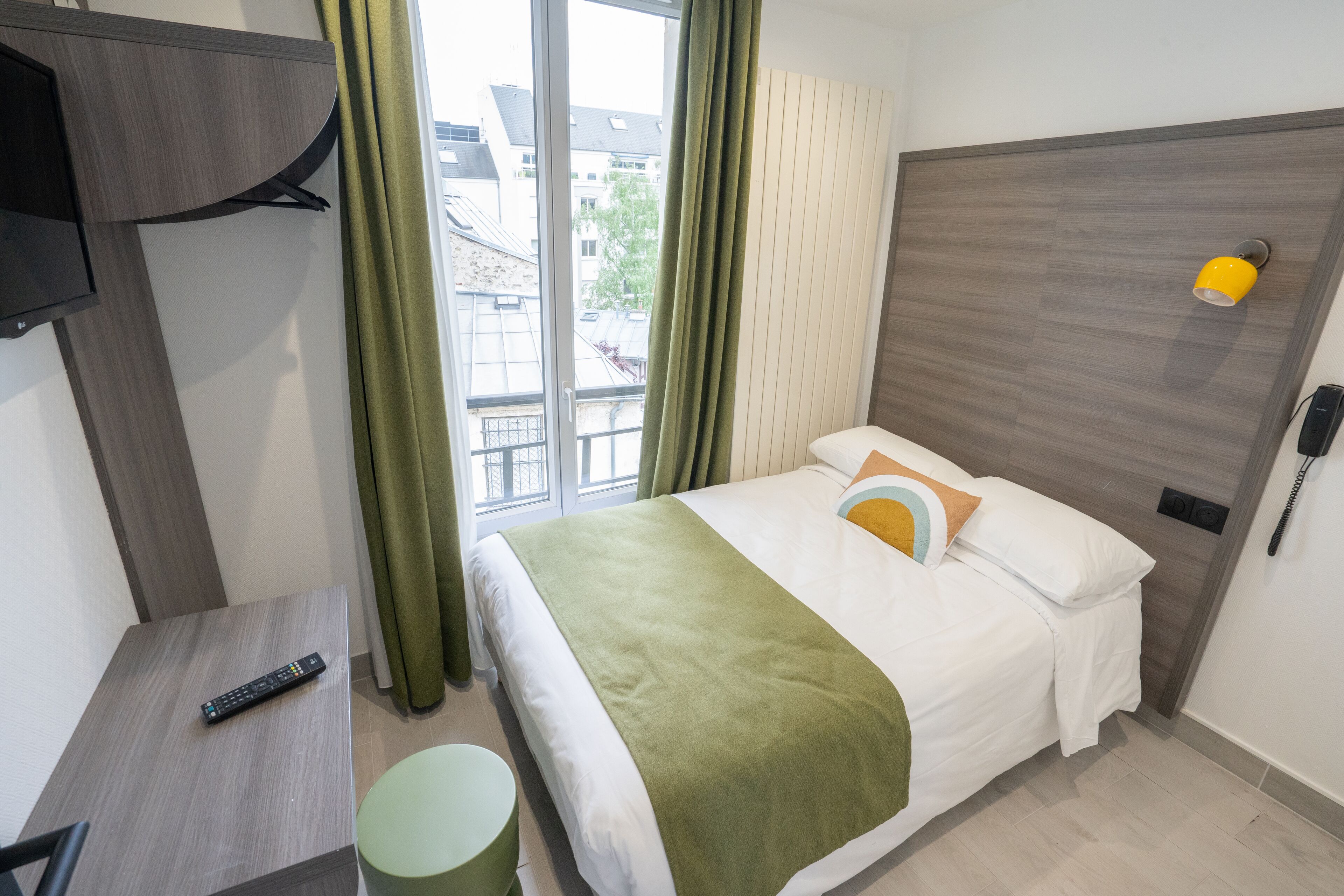 A hotel room with a bed, a TV mounted on the wall, a bedside table with a remote control, and a window with curtains.