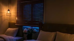 A bedroom with a bed, two bedside tables, a lamp, and a window with blinds. - Hi Jun Guesthouse (Seoul)