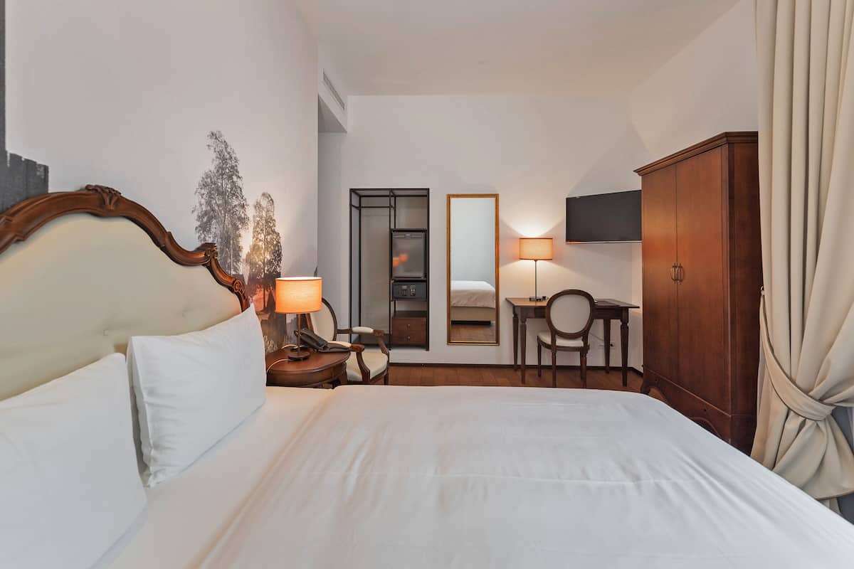Ein Hotelzimmer mit einem großen Bett, einem Nachttisch mit Lampe, einem Schreibtisch mit Stuhl, einem Kleiderschrank und einem Spiegel an der Wand.