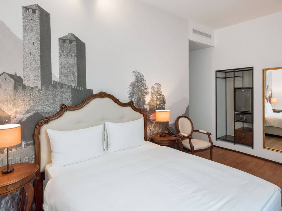Una habitación de hotel con una cama grande, dos mesitas de noche con lámparas, una silla, un espejo y un mural en la pared con un castillo.