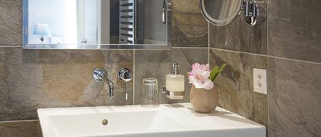 Un baño con lavamanos blanco, espejo, dispensador de jabón de vidrio y un florero con flores.