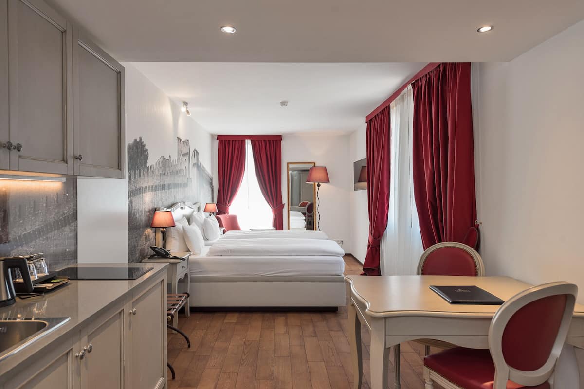 Ein Hotelzimmer mit Bett, Schreibtisch, Stuhl, einer kleinen Küchenzeile und einem durch eine Tür einsehbaren Badezimmer.
