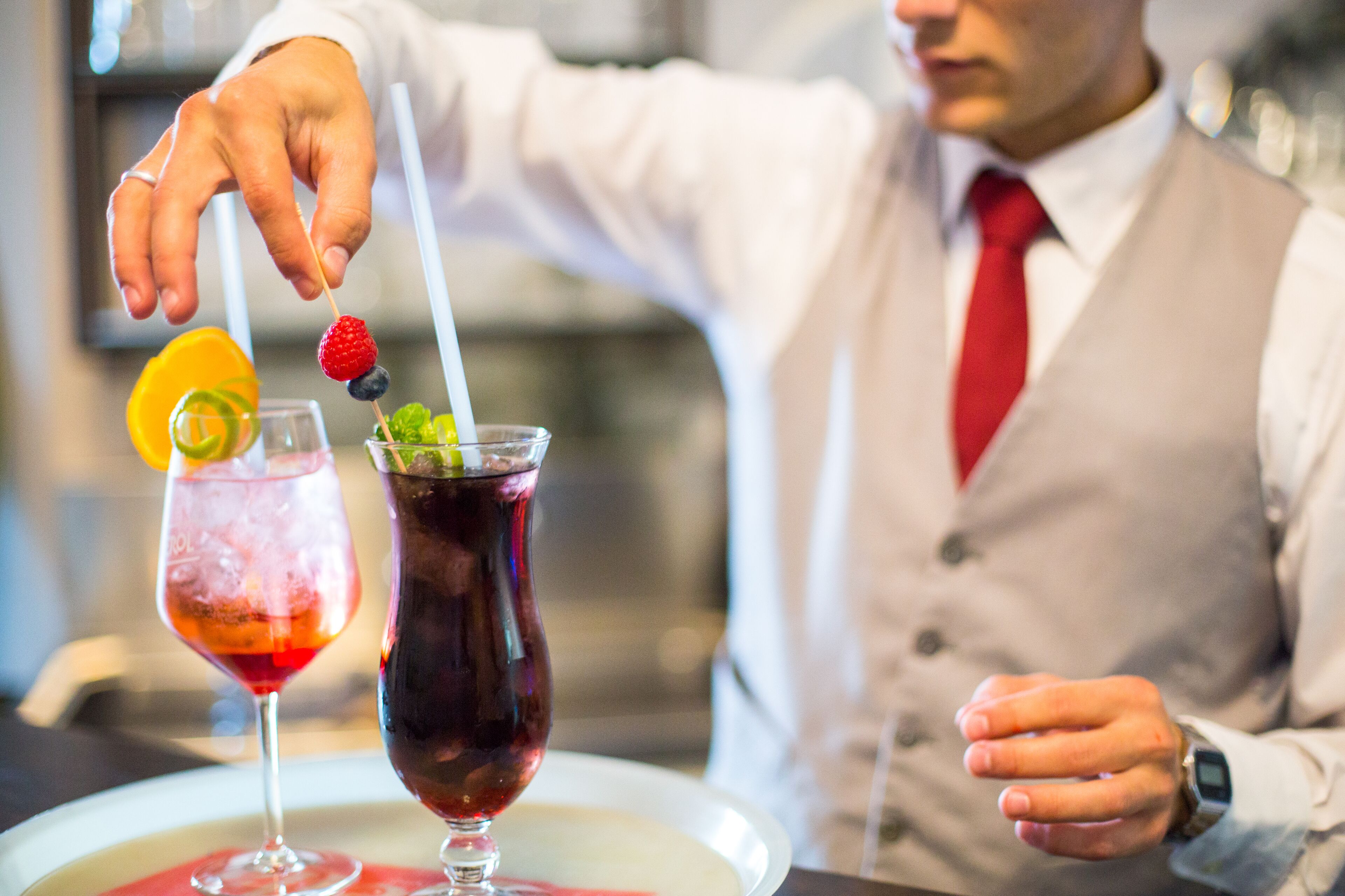 A bartender preparing drinks with a tray of two cocktails, one with a red drink and the other with a dark purple drink, garnished with fruits and a straw.
