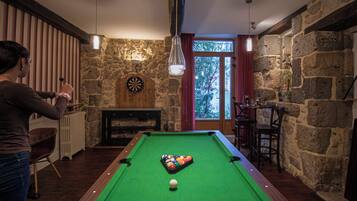 Una mesa de billar en una habitación con paredes de piedra, un dardo y un ventanal con vista a árboles.