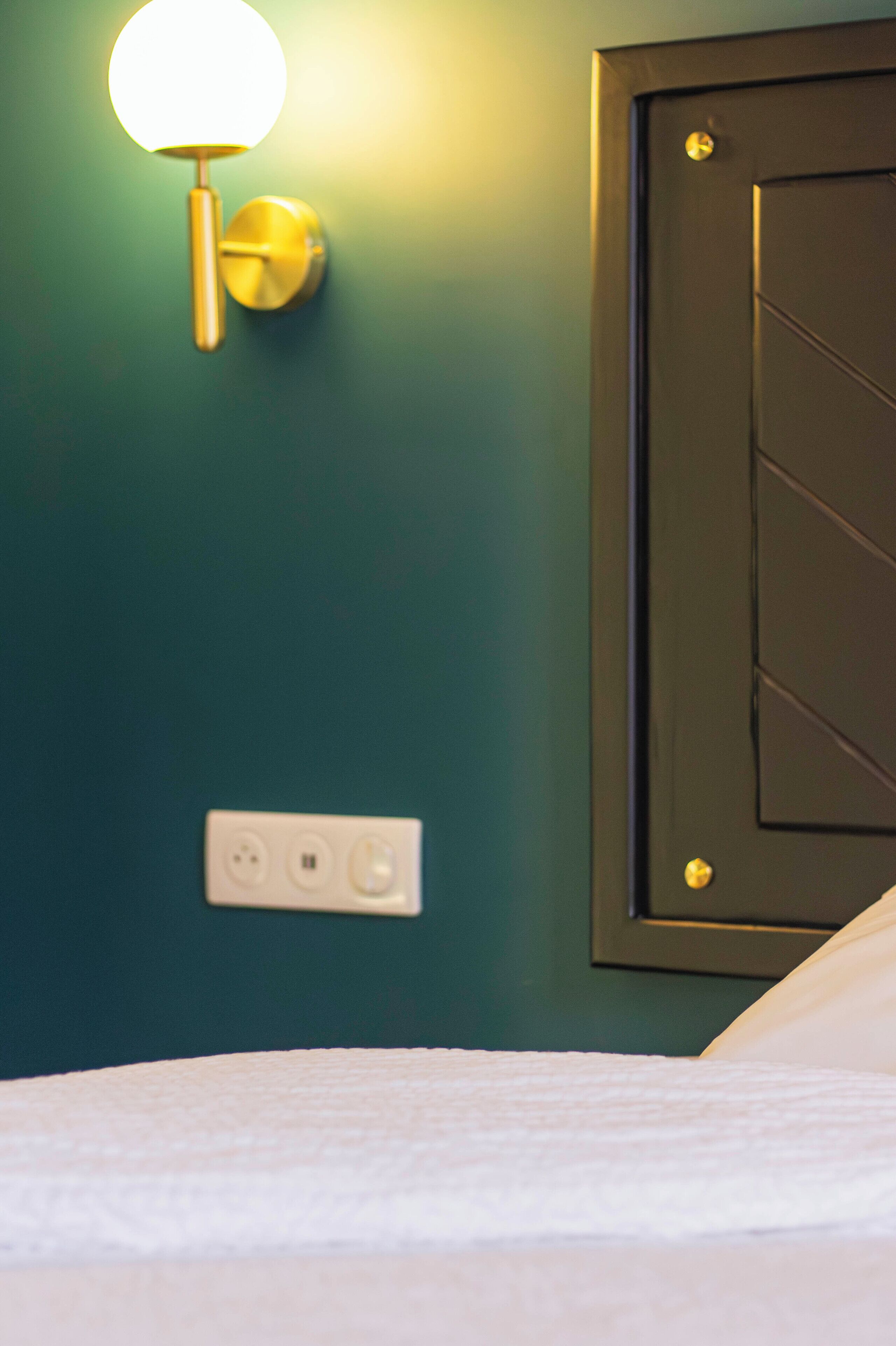 A hotel room with a teal wall, a wall-mounted light fixture, a door with a handle, and a power outlet.