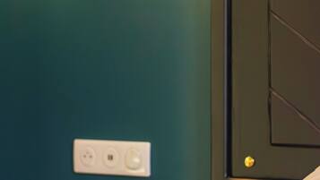 A hotel room with a teal wall, a wall-mounted light fixture, a door with a handle, and a power outlet.
