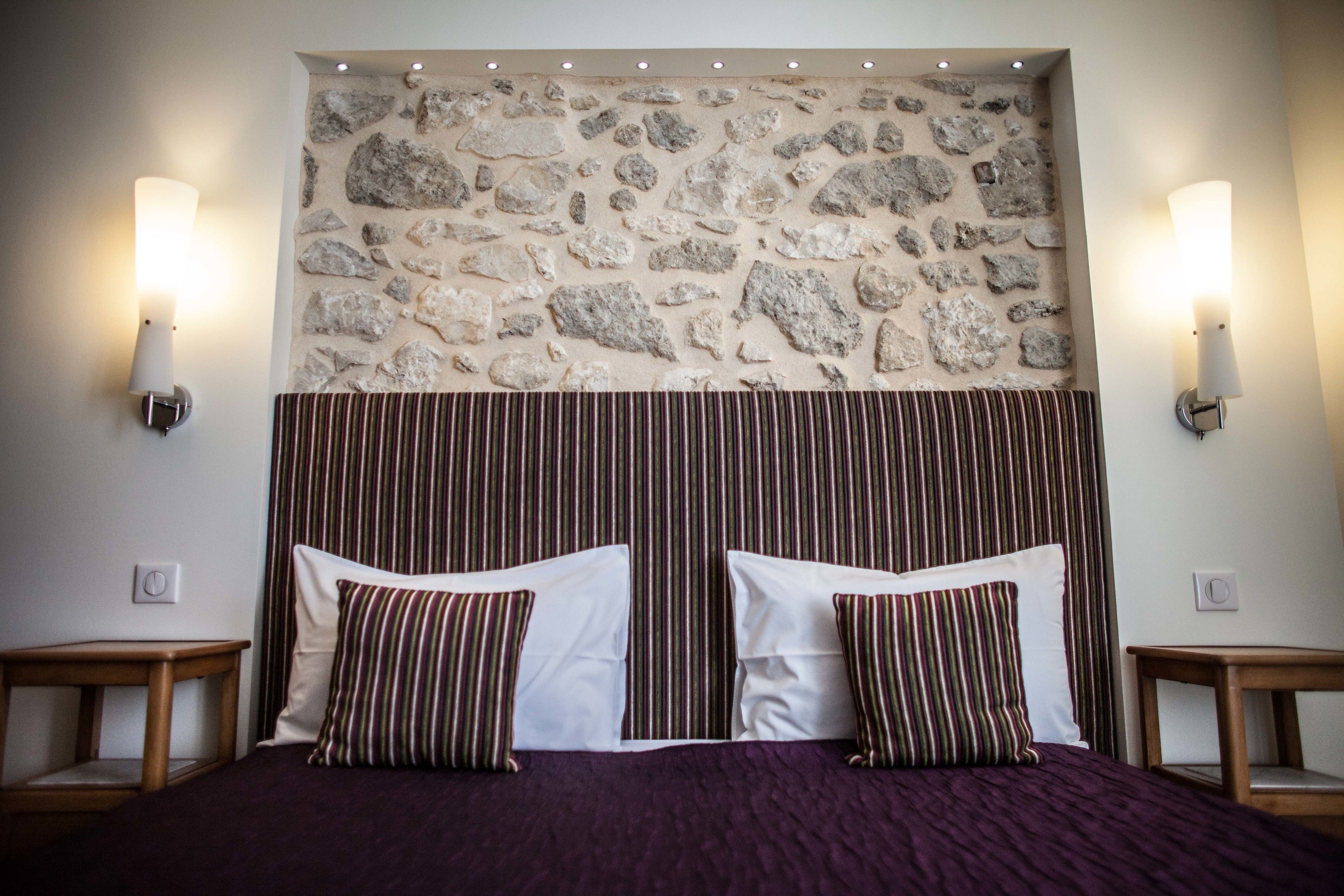 Habitación de hotel con pared de piedra, dos lámparas de noche, una cama con almohadas blancas y a rayas, y un cabecero con rayas verticales.