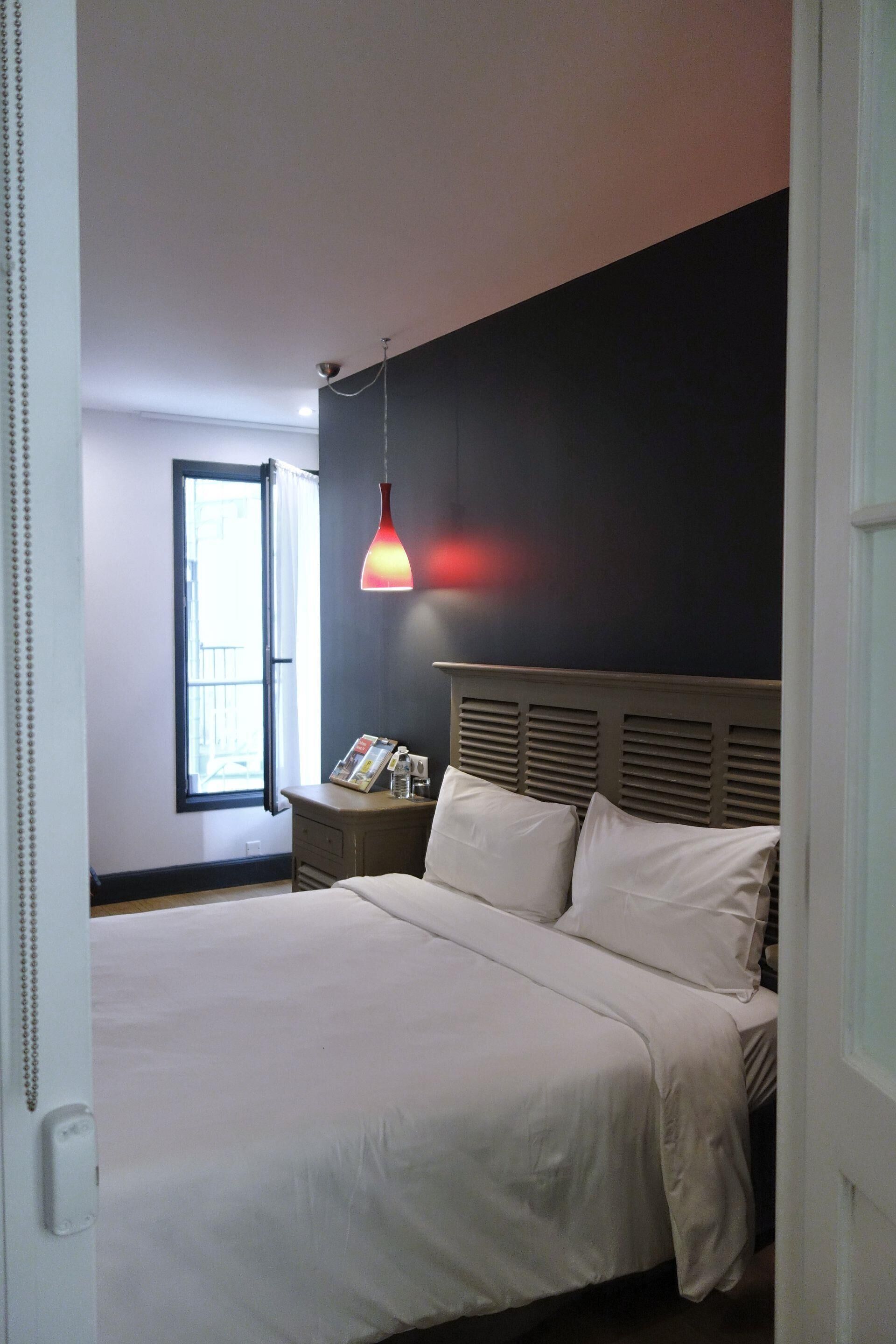 Quarto com cama, mesa de cabeceira, luminária e janela com cortinas.