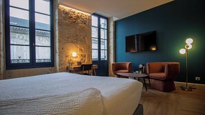 Habitación de hotel con una cama grande, un escritorio con lámpara, dos sillas, un televisor montado en la pared y una pared de piedra.