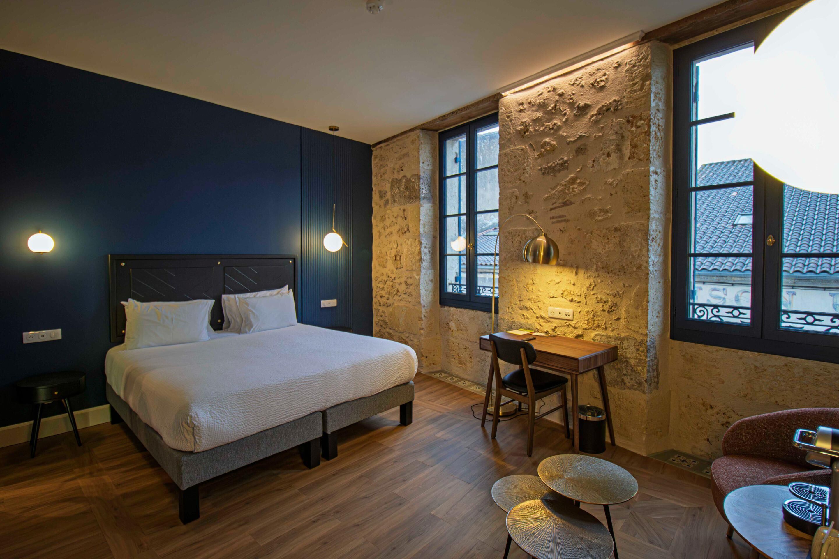 A bedroom with a bed, desk, chair, and a view of buildings through the window.
