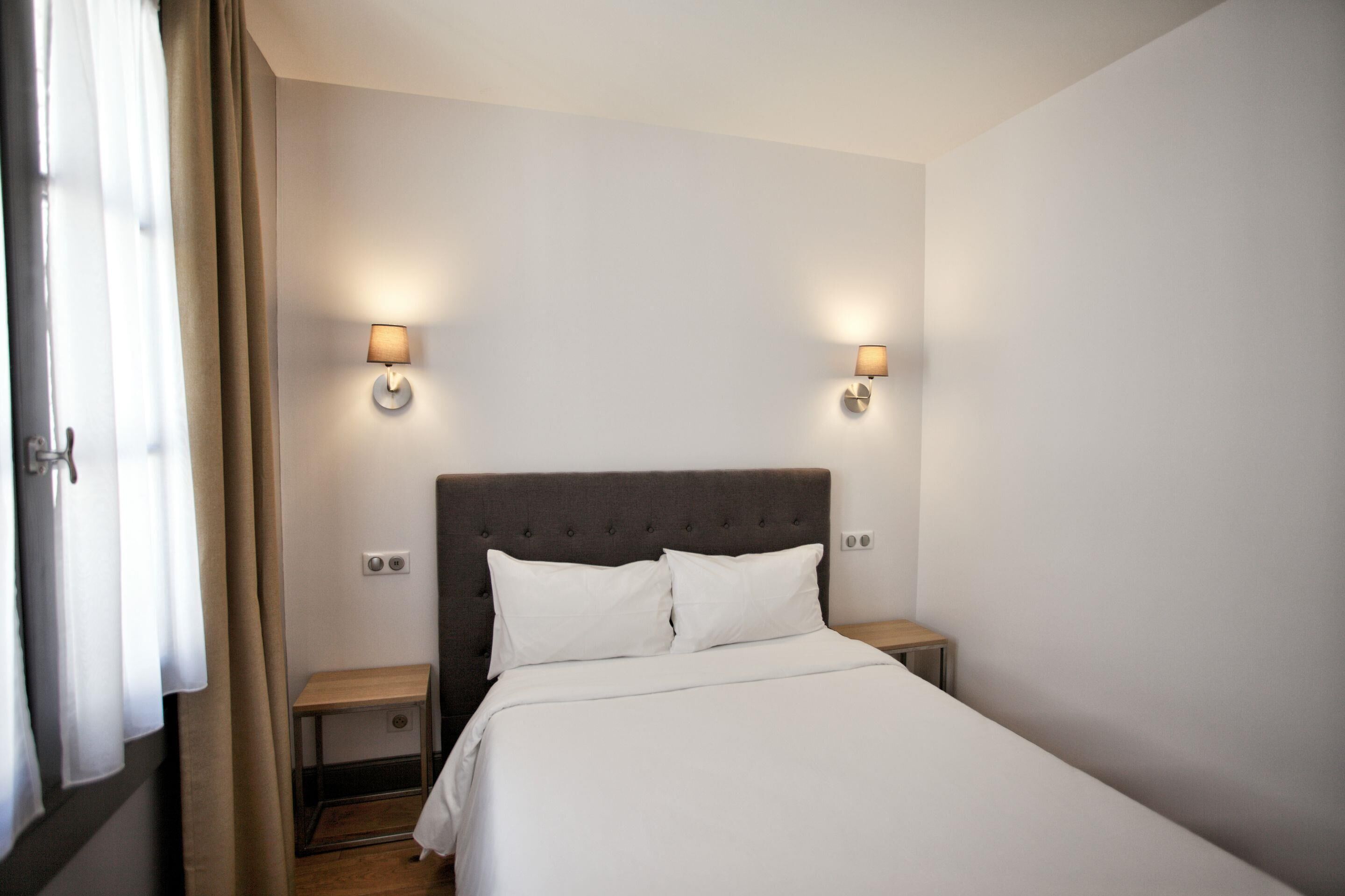 A hotel room with a bed, two bedside tables, two wall-mounted lamps, and a window with curtains.