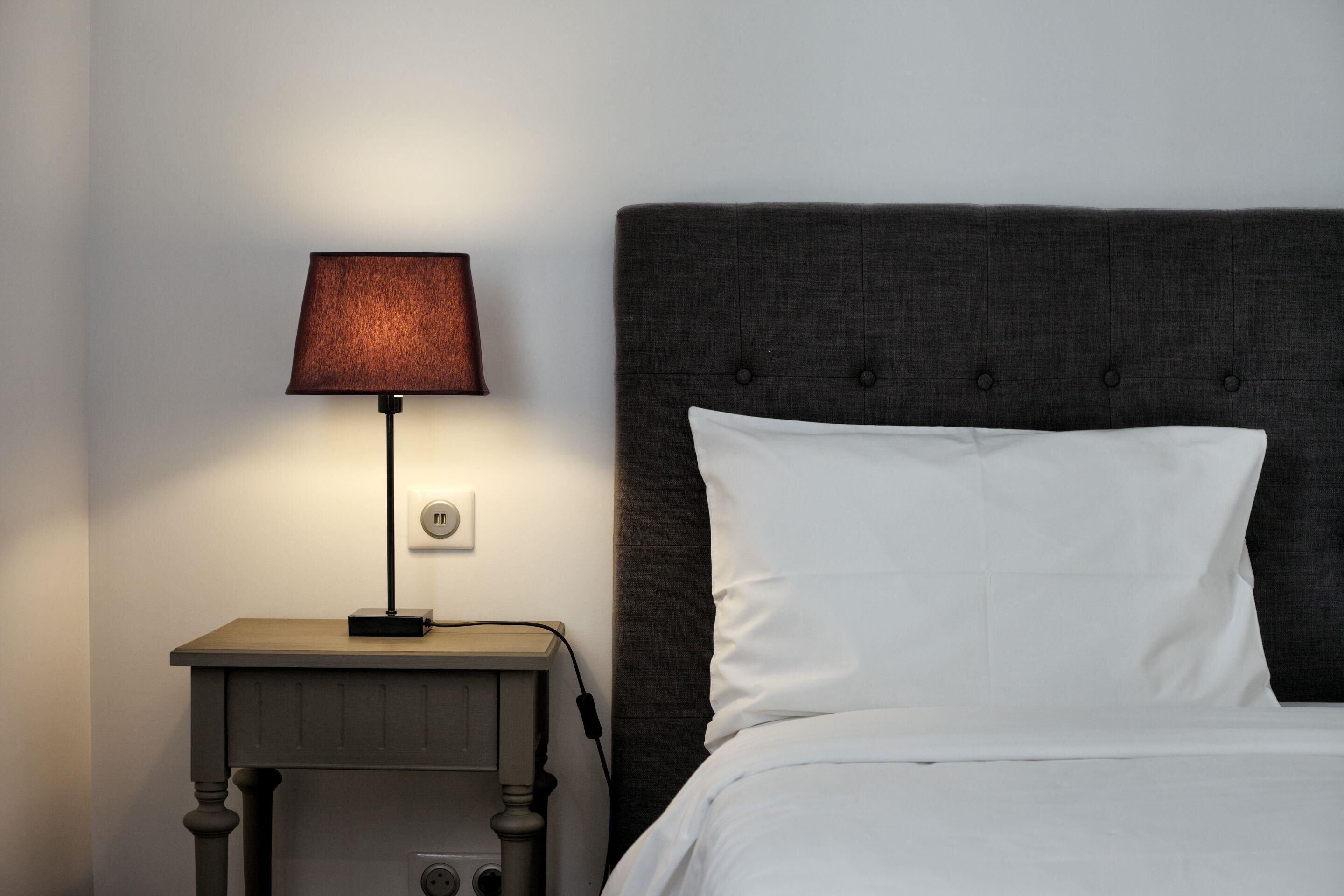 A neatly made bed with a dark headboard, a bedside lamp, and a power outlet on a wooden nightstand.